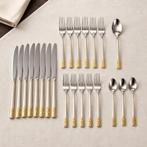 May include: A complete silver and gold-toned flatware set arranged on a beige tablecloth. The set includes knives, forks, and spoons. The handles have a decorative gold design. The arrangement suggests a formal dining setting.