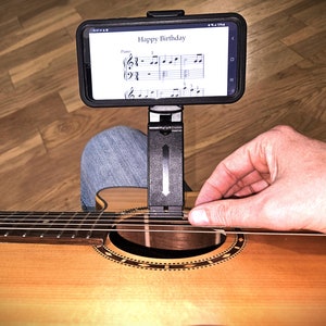 May include: A person playing a brown acoustic guitar with a phone attached to a stand displaying sheet music for "Happy Birthday".