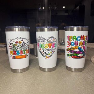 May include: Three white insulated tumblers with silver rims. Each tumbler features a different design: one with floral and pencil graphics and the word "MAESTRA", another with a heart-shaped design and the word "MAESTRA", and the last with the text "TEACHER YOU ARE" and crayon graphics.