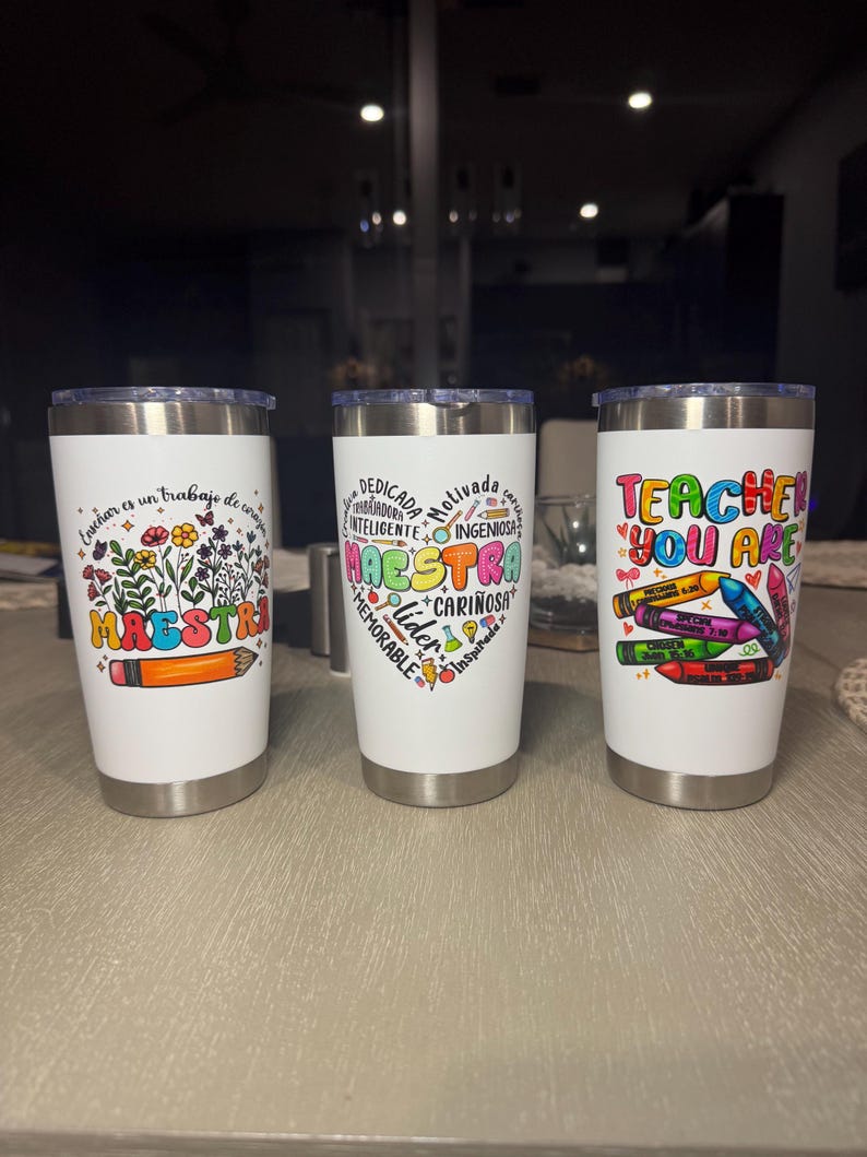 May include: Three white insulated tumblers with silver rims. Each tumbler features a different design: one with floral and pencil graphics and the word "MAESTRA", another with a heart-shaped design and the word "MAESTRA", and the last with the text "TEACHER YOU ARE" and crayon graphics.