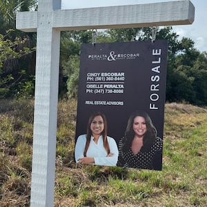 May include: A white wooden for sale sign with a black rectangular sign hanging from it. The black sign has the names and photos of two real estate agents, along with their contact information. The word "FORSALE" is printed vertically on the right side.