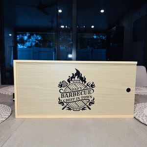 May include: A rectangular wooden box with a light wood grain finish. The box features a black graphic that reads "DAD'S BARBECUE BEST IN TOWN" with grilling tools and flames. A small black hole is on the right side.