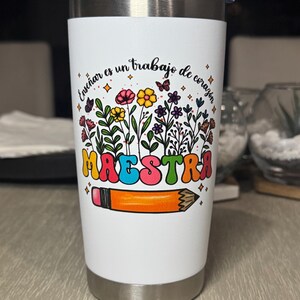 May include: A white insulated tumbler with a silver lid. The tumbler features a colorful floral design with the word "MAESTRA" in large, retro-style letters, and a pencil illustration. The text "Ense&ntilde;ar es un trabajo de coraz&oacute;n" is also present.