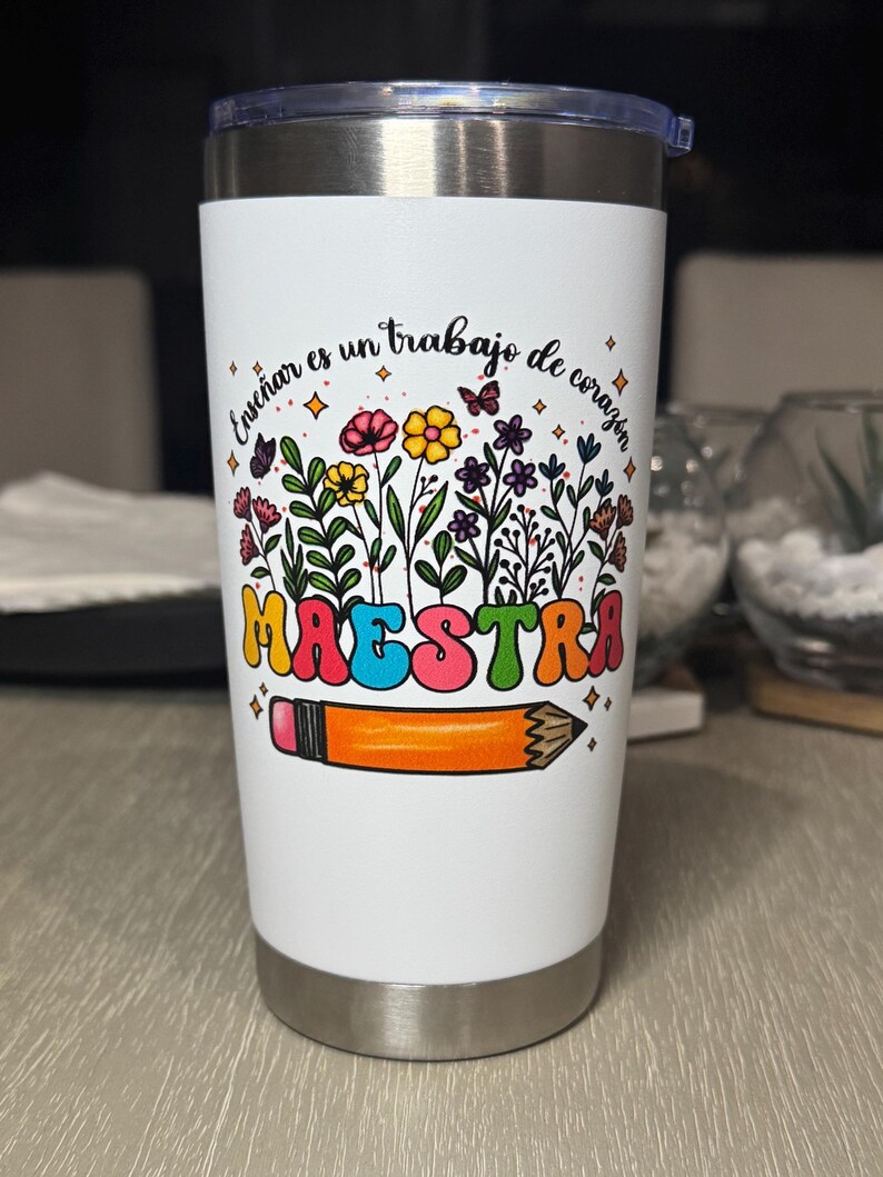 May include: A white insulated tumbler with a silver lid. The tumbler features a colorful floral design with the word "MAESTRA" in large, retro-style letters, and a pencil illustration. The text "Ense&ntilde;ar es un trabajo de coraz&oacute;n" is also present.