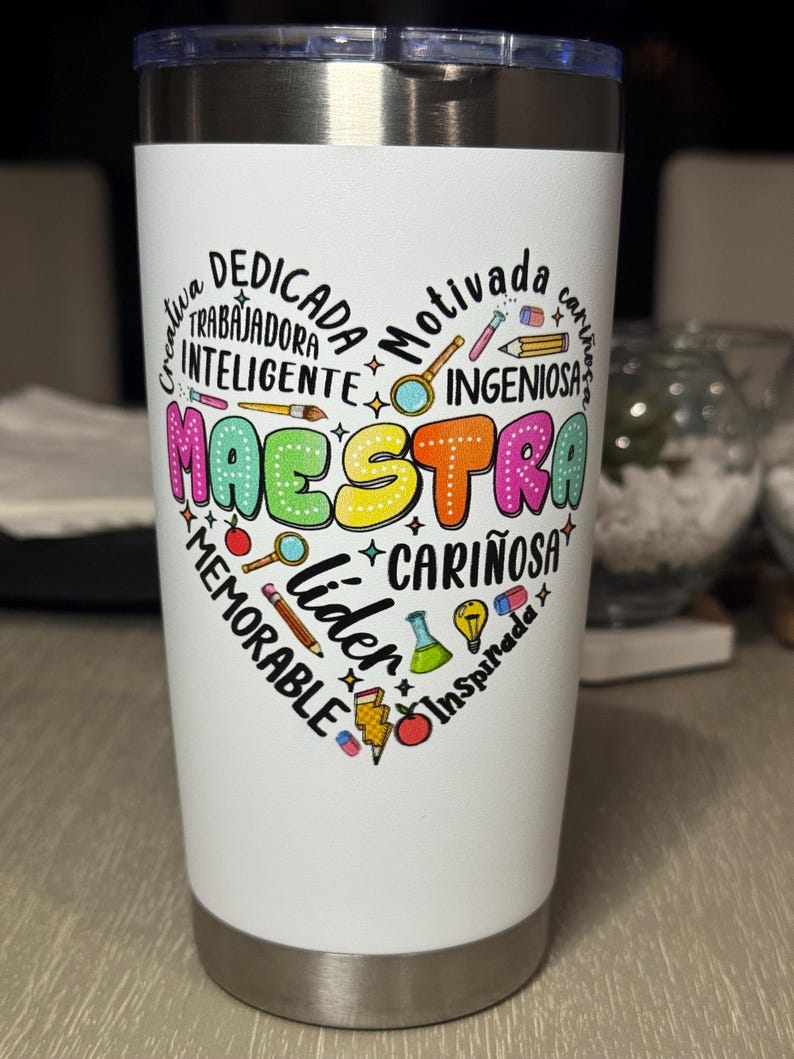 May include: A white insulated tumbler with a silver rim and lid. The tumbler features a heart-shaped design with the word "MAESTRA" in colorful letters, surrounded by words like "creative," "dedicated," and "leader."