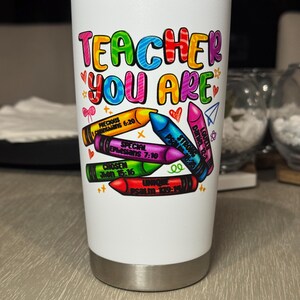 May include: A white insulated tumbler with a colorful design. The text "TEACHER YOU ARE" is displayed in rainbow colors. Below the text are crayon illustrations with various inspirational words and Bible verses. The tumbler has a silver base and a silver lid.