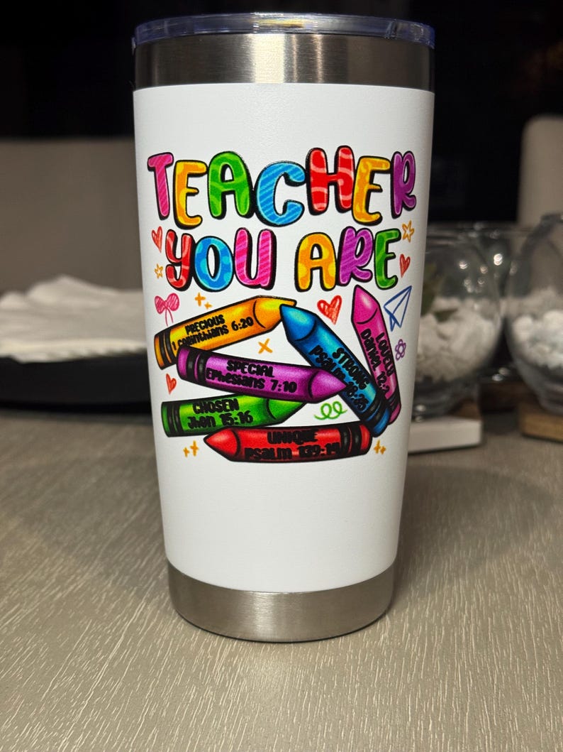 May include: A white insulated tumbler with a colorful design. The text "TEACHER YOU ARE" is displayed in rainbow colors. Below the text are crayon illustrations with various inspirational words and Bible verses. The tumbler has a silver base and a silver lid.