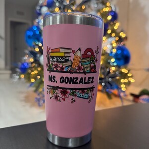 May include: A pink insulated tumbler with a silver rim and base. The tumbler features a colorful graphic of books, a pencil, and a coffee cup, with the text "MS. GONZALEZ" in black. The design is framed by floral accents.
