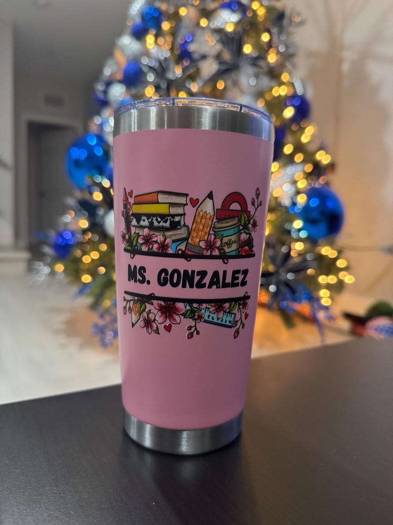 May include: A pink insulated tumbler with a silver rim and base. The tumbler features a colorful graphic of books, a pencil, and a coffee cup, with the text "MS. GONZALEZ" in black. The design is framed by floral accents.