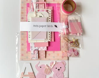 Könnte beinhalten: Eine Sammlung von rosa und weißen Schreibwaren, darunter gemustertes Papier, Etiketten, Washi-Tape und Verzierungen. Das Set zeichnet sich durch eine Vielzahl von Texturen und Designs aus, mit einem Baby-Thema, einschließlich eines Elefanten und eines Teddybären.