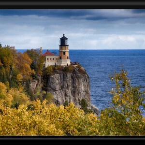 May include: A lighthouse stands on a rocky cliff overlooking a blue body of water. The lighthouse is white with a black roof and a black lantern. The cliff is covered in trees with yellow leaves. The sky is cloudy and blue.