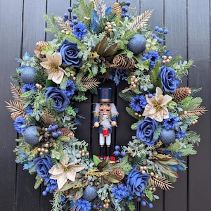 May include: A circular festive wreath featuring deep blue roses, champagne-coloured poinsettias, pinecones, blue berries, and green pine needles. A nutcracker figure with a blue uniform and red drum is positioned in the centre of the wreath.