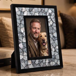 Puede incluir: Un marco de fotos rectangular con borde negro y una incrustación interior de piezas de concha iridiscentes estilo mosaico en tonos blanco, gris y plata. El marco contiene una fotografía de una persona con un perro en orientación vertical.