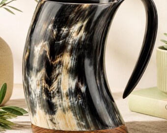 May include: A drinking vessel crafted from a polished animal horn with a curved silhouette and a dark, marbled pattern of black, cream, and brown tones. The mug features a sturdy, integrated handle and a flat, circular wooden base.