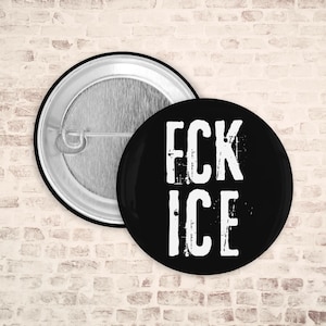 Pode incluir: Um botão preto com o texto "FCK ICE" em branco, com letras desgastadas. O botão é redondo e tem um alfinete de metal na parte de trás. O fundo é uma parede de tijolos.
