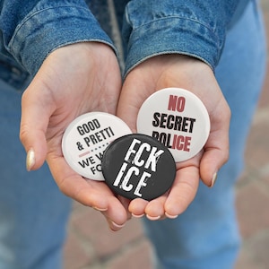 May include: Three round buttons with text are held in open hands. One button is black with white text that reads "FCK ICE". Another is white with black text that reads "NO SECRET POLICE". The third is white with pink text that reads "GOOD & PRETTI".