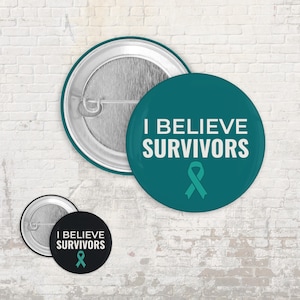 Könnte beinhalten: Runde Ansteckbuttons in Türkis und Schwarz mit dem weißen Text "I BELIEVE SURVIVORS" und einem türkisfarbenen Schleifensymbol. Die Buttons haben einen Metall-Pin-Rücken. Der türkisfarbene Button ist größer.