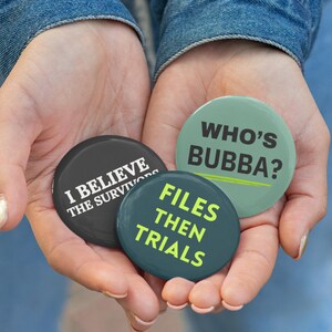 Könnte beinhalten: Drei runde Buttons mit Text werden in offenen Händen gehalten. Die Buttons sind schwarz, blaugrün und grün. Der schwarze Button lautet "I BELIEVE THE SURVIVORS" in Weiß. Der blaugrüne Button lautet "FILES THEN TRIALS" in Gelb. Der grüne Button lautet "WHO'S BUBBA?" in Schwarz.