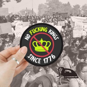 May include: A round, black patch with the text "NO FUCKING KINGS" and "SINCE 1776" in white. A yellow crown is crossed out with a red line. The patch is held in front of a crowd of people.