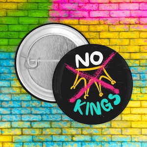 May include: Two round buttons. One is silver, the other is black with the words "NO KINGS" in white and turquoise, with a crossed-out crown in pink and yellow. The background is a colorful brick wall.