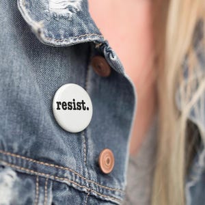 Könnte beinhalten: Ein weißer Ansteckbutton mit dem Wort "resist." in schwarzer, fetter Schrift. Der Button ist an einer blauen Jeansjacke befestigt. Die Jacke hat kupferfarbene Knöpfe und einen Used-Look.