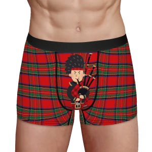 May include: Boxer briefs with a red, green, and black plaid pattern. The briefs feature a cartoon illustration of a bagpiper in traditional Scottish attire. The waistband is black.