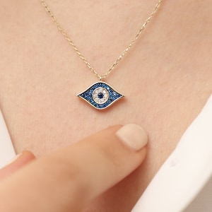 Puede incluir: Un collar de cadena de oro con un colgante de ojo turco azul y blanco. El colgante está engastado con pequeñas piedras blancas y tiene un ojo de esmalte azul.