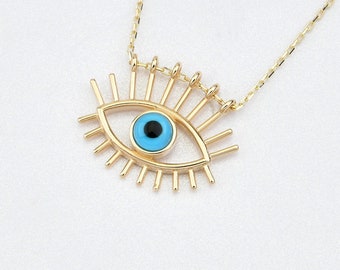 May include: Gold necklace featuring a blue evil eye pendant. The pendant is a stylised eye with a blue centre and gold lashes.