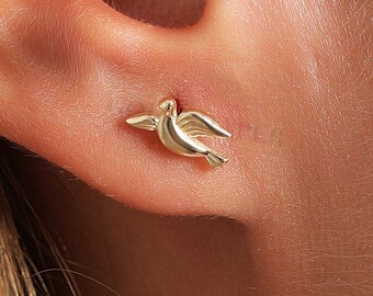 May include: A gold dove-shaped stud earring. The earring is small and delicate, and it is shown in the ear of a person.