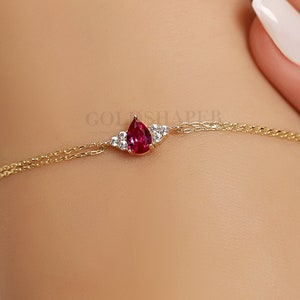 Könnte beinhalten: Ein zartes Armband aus einer goldenen Kette mit einem tropfenförmigen roten Edelstein, der von kleineren klaren Edelsteinen umgeben ist.