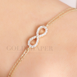 Puede incluir: Un brazalete de oro con un símbolo de infinito con pequeñas piedras transparentes. El brazalete está en la muñeca de una persona.