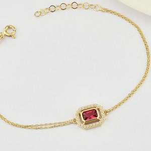 Puede incluir: Un brazalete de cadena dorada con una piedra preciosa roja engastada en un marco cuadrado con piedras transparentes alrededor del borde.
