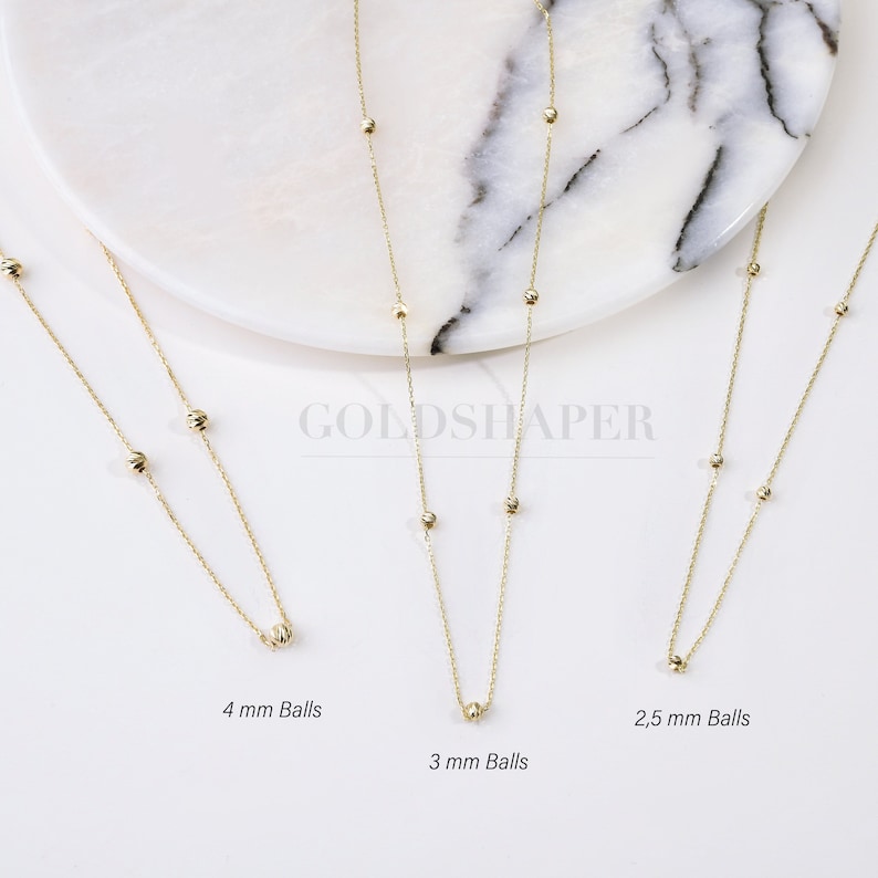 May include: Three gold necklaces with different sized balls. The necklaces are on a white marble surface. The balls are 2.5 mm, 3 mm, and 4 mm in diameter.