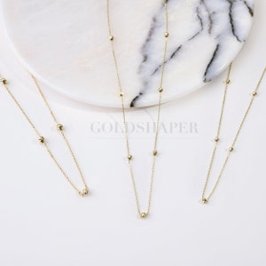 May include: Three gold necklaces with small round beads. The necklaces are on a white marble surface.