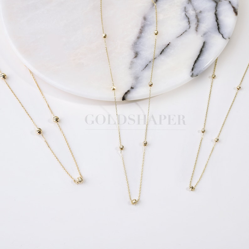 May include: Three gold necklaces with small round beads. The necklaces are on a white marble surface.