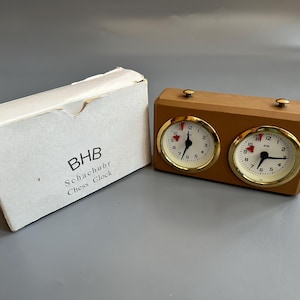 Könnte beinhalten: Eine Vintage-Schachuhr mit einem hellbraunen Holzgehäuse und zwei runden Zifferblättern mit goldfarbenen Lünetten. Daneben liegt eine weiße Schachtel mit der Aufschrift "BHB Schachur Chess Clock".