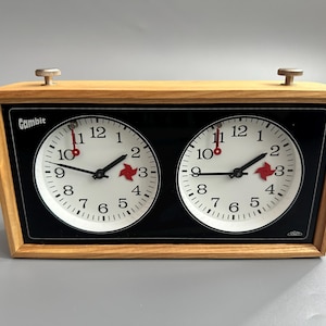 Puede incluir: Reloj de ajedrez vintage con dos esferas blancas y un marco de madera. Las manecillas son negras y un símbolo rojo de ajedrez está a las 3 en punto. La palabra "Gambit" está impresa en la parte superior izquierda. Mide el tiempo de juego.