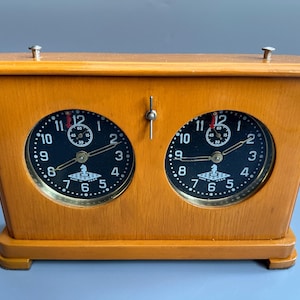 May include: A vintage wooden chess clock with two black clock faces. Each clock face has white numerals and hands, with a small chess piece logo at the bottom. The wooden case is light brown.