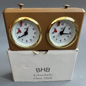 May include: A brown wooden chess clock with two round clock faces. Each clock face has a gold-coloured bezel, white background, and black hands. The clock is labelled "BHB Schachur Chess Clock" on the box below.