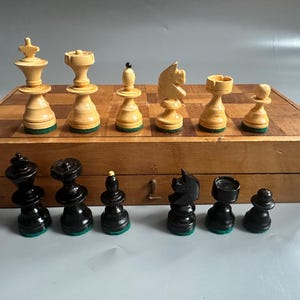 以下が含まれることがあります： 黒とクリーム色の駒が入った、木製のヴィンテージチェスセット。 チェスの駒は木製のチェス盤に配置され、駒は初期位置にあります。 セットは木製の箱に収納されています。