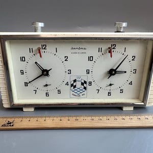 May include: Vintage chess clock with a cream-colored face and a silver frame. The clock has two dials with black hands and red second hands. The text "lantaz MADE IN USSR" is visible. The clock also features the FISU logo and the text "ODESSA 90".