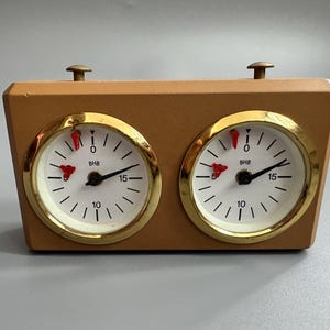 Può includere: Un orologio da scacchi vintage con una cassa in legno marrone chiaro e due quadranti circolari. Ogni quadrante ha una lunetta color oro, un quadrante bianco con numeri neri e un indicatore rosso. L'orologio ha due manopole color ottone sulla parte superiore.