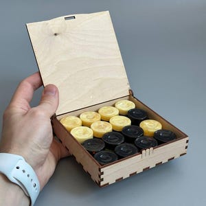 May include: A square light-colored wooden box with an open hinged lid containing a set of round game pieces. The game pieces are arranged in rows and consist of twelve pale yellow circular tokens and twelve dark black circular tokens.