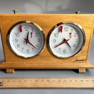 May include: A wooden chess clock with two round clock faces. Each clock face has a white background, black numbers, and red hands. The clock is made of light-colored wood and has a gold-colored trim around the clock faces.