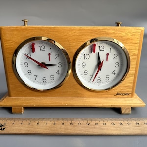 May include: A wooden chess clock with two round faces, each with a white dial, black numerals, and red hands. The clock has a gold-colored bezel and is set on a wooden base. A ruler is placed below the clock.