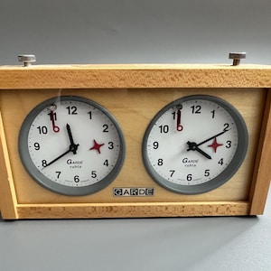 Può includere: Un orologio da scacchi in legno con due quadranti circolari. Ogni quadrante ha uno sfondo bianco, lancette nere e indicatori rossi a forma di stella. L'orologio è etichettato "GARDE ruhla".