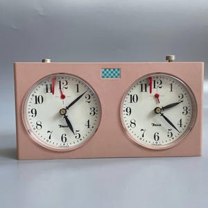 May include: A vintage pink chess clock with two round clock faces. Each clock face has black hands, white numbers, and a red second hand. A small checkered pattern is above the clock faces.