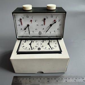 Peut inclure: Une horloge d'échecs vintage avec un cadre noir, des cadrans blancs et des aiguilles des secondes rouges. L'horloge a deux boutons blancs sur le dessus et est présentée sur sa boîte blanche d'origine. Le cadran affiche les chiffres de 1 à 12.