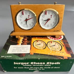 May include: A vintage Jerger chess clock with two round analog clocks. The clock has a wooden frame and gold-colored bezels. The clock face is white with black numbers and red hands. The clock sits on a green and brown box with text.