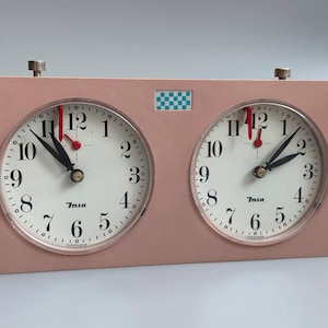 May include: A rectangular pink analogue chess clock with two circular white faces featuring black numerals and the brand name Insa. Each clock face has black hour and minute hands, a red second hand, and a small checkered logo centred above.